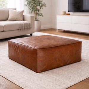 May include: A large, square, brown leather ottoman. The ottoman is a warm, medium brown color and appears to be made of a soft, textured leather. It sits on a light-colored rug in a living room setting.