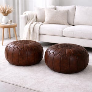 May include: Two brown leather poufs with geometric stitching. The poufs are round and sit on a white rug. A white sofa with pillows and a blanket is in the background. A small wooden table with a vase of dried flowers is also visible.