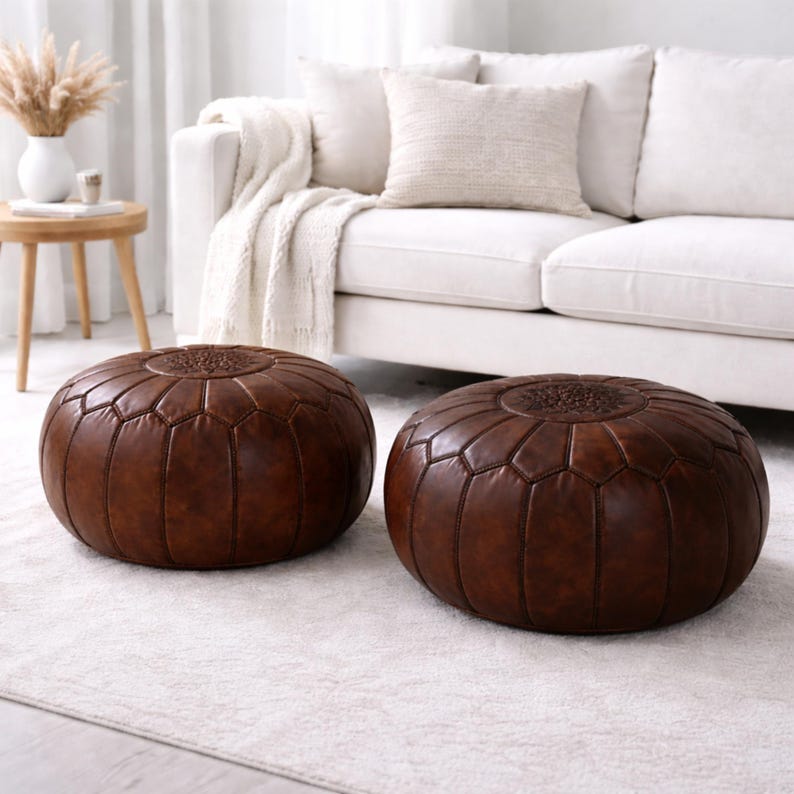 May include: Two brown leather poufs with geometric stitching. The poufs are round and sit on a white rug. A white sofa with pillows and a blanket is in the background. A small wooden table with a vase of dried flowers is also visible.
