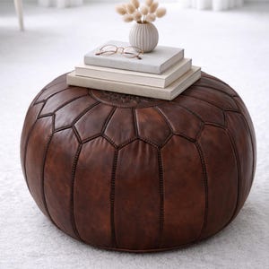 May include: A round, brown leather ottoman with a geometric pattern. The ottoman is topped with a stack of books, a small vase with dried flowers, and a pair of eyeglasses. The furniture piece is a decorative and functional item for a living room or bedroom.