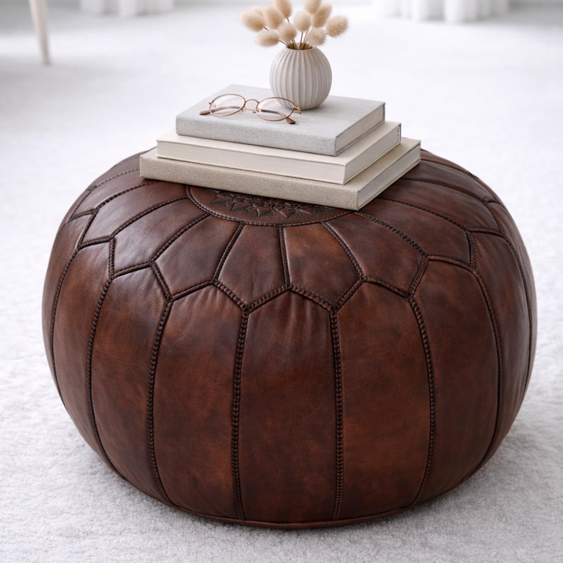 May include: A round, brown leather ottoman with a geometric pattern. The ottoman is topped with a stack of books, a small vase with dried flowers, and a pair of eyeglasses. The furniture piece is a decorative and functional item for a living room or bedroom.
