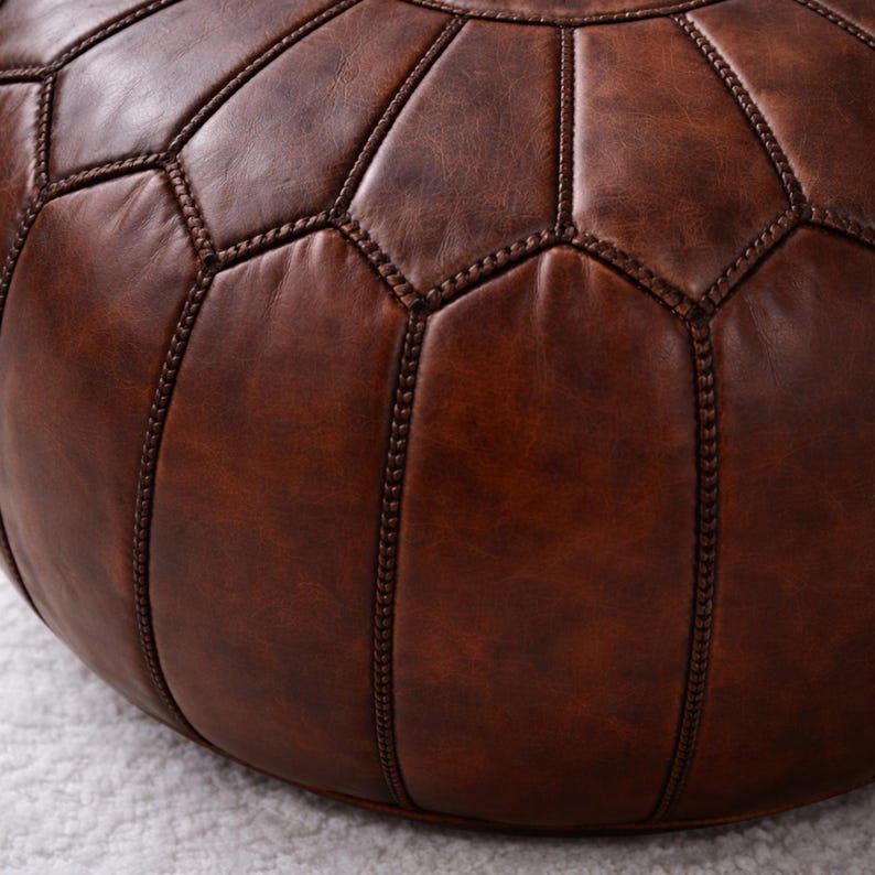 May include: A brown leather ottoman with a geometric pattern. The round pouf is made of stitched panels, creating a textured surface. The color is a rich, warm brown, suitable for a living room or bedroom.