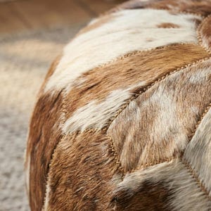 May include: Close-up of a round, patchwork ottoman. The ottoman features a pattern of brown, tan, and white cowhide pieces stitched together with a tan thread. The texture of the cowhide is visible.