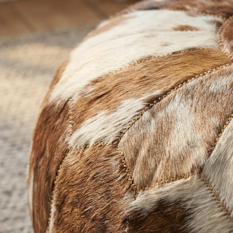 May include: Close-up of a round, patchwork ottoman. The ottoman features a pattern of brown, tan, and white cowhide pieces stitched together with a tan thread. The texture of the cowhide is visible.