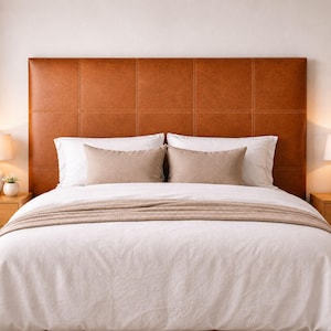 May include: A rectangular headboard upholstered in tan leather with a grid-patterned stitched design. The bed features a white duvet cover, two white square pillows, two rectangular beige accent pillows, and a beige throw blanket.