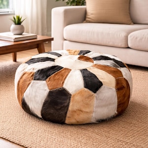Può includere: Un pouf rotondo patchwork con un motivo geometrico. Il pouf presenta un mix di colori tra cui bianco, nero, marrone e beige. È posizionato su un tappeto di colore neutro, con un divano e un tavolino sullo sfondo.