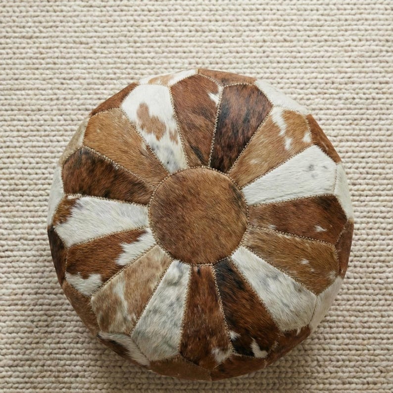 May include: Round ottoman with a patchwork design made from cowhide. The ottoman features a mix of brown, white, and tan patches arranged in a radial pattern. The center is a solid brown circle.