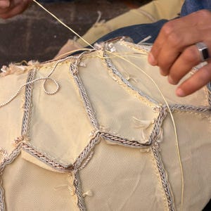 May include: Close-up of a craft project in progress, featuring a cream-colored fabric with a geometric pattern. The edges are trimmed with a silver and brown braid. A person is sewing the fabric with a light yellow thread.