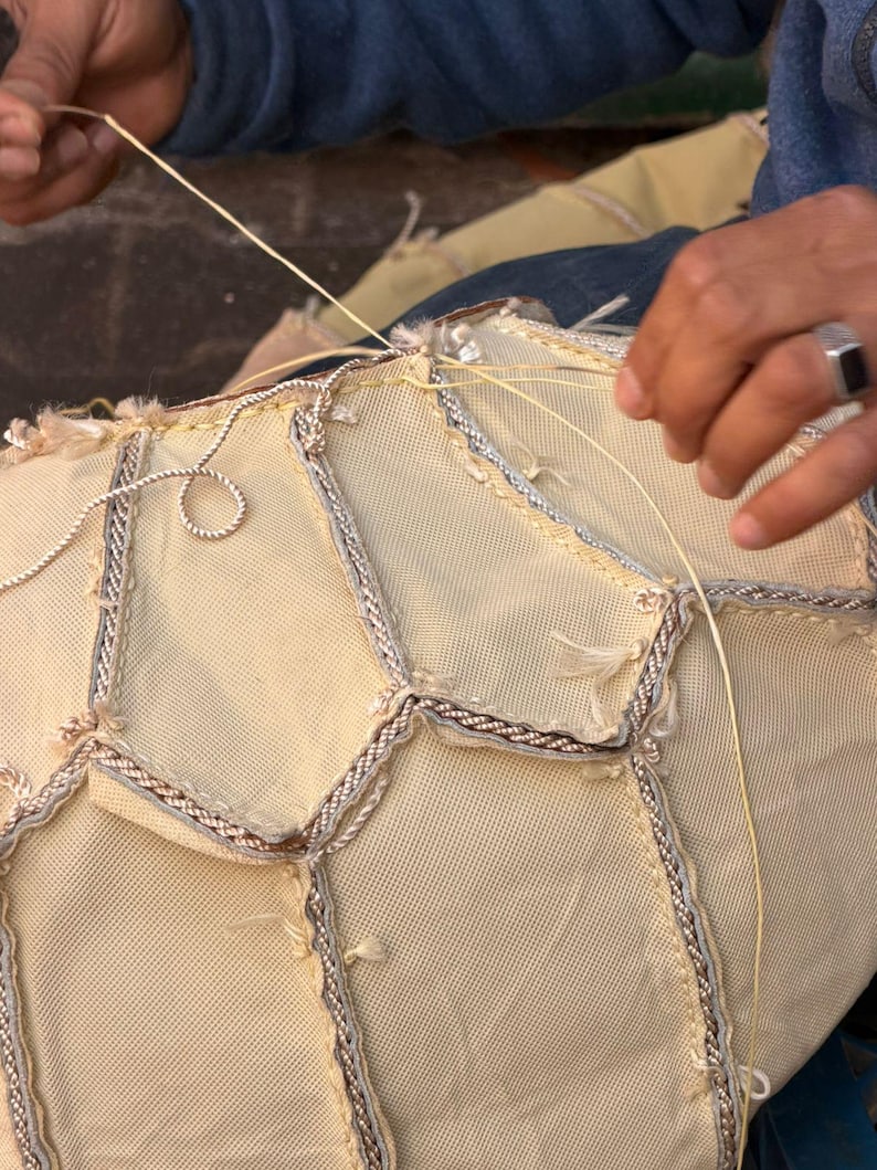 May include: Close-up of a craft project in progress, featuring a cream-colored fabric with a geometric pattern. The edges are trimmed with a silver and brown braid. A person is sewing the fabric with a light yellow thread.