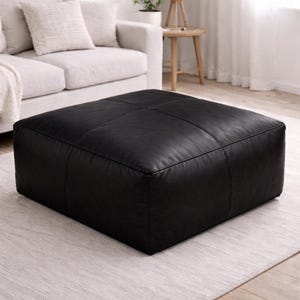 Peut inclure: Un pouf carré en cuir noir avec des coutures décoratives. Le pouf est posé sur un tapis clair, avec un canapé en arrière-plan. Un meuble polyvalent pour le salon ou la chambre.