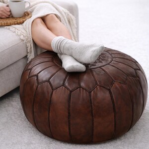 May include: A round, brown leather ottoman with a geometric pattern. The ottoman is a dark, rich brown color and has visible stitching. The top of the ottoman has a decorative floral design. The ottoman is on a light gray carpet.