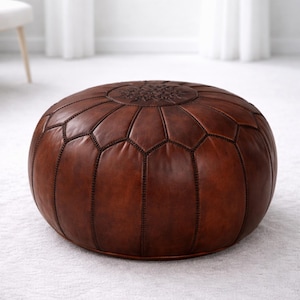 May include: A round, brown leather ottoman with a geometric pattern and decorative stitching. The top features a circular embossed design. The ottoman is placed on a white carpet, offering a comfortable and stylish seating option.