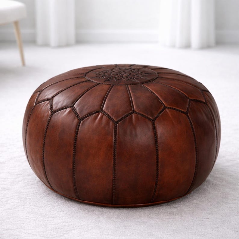 May include: A round, brown leather ottoman with a geometric pattern and decorative stitching. The top features a circular embossed design. The ottoman is placed on a white carpet, offering a comfortable and stylish seating option.