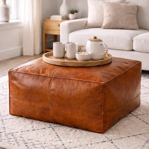May include: A large, square, brown leather ottoman in a living room setting. A wooden tray holds white mugs, a bowl of snacks, and a white teapot. The ottoman is placed on a patterned rug.
