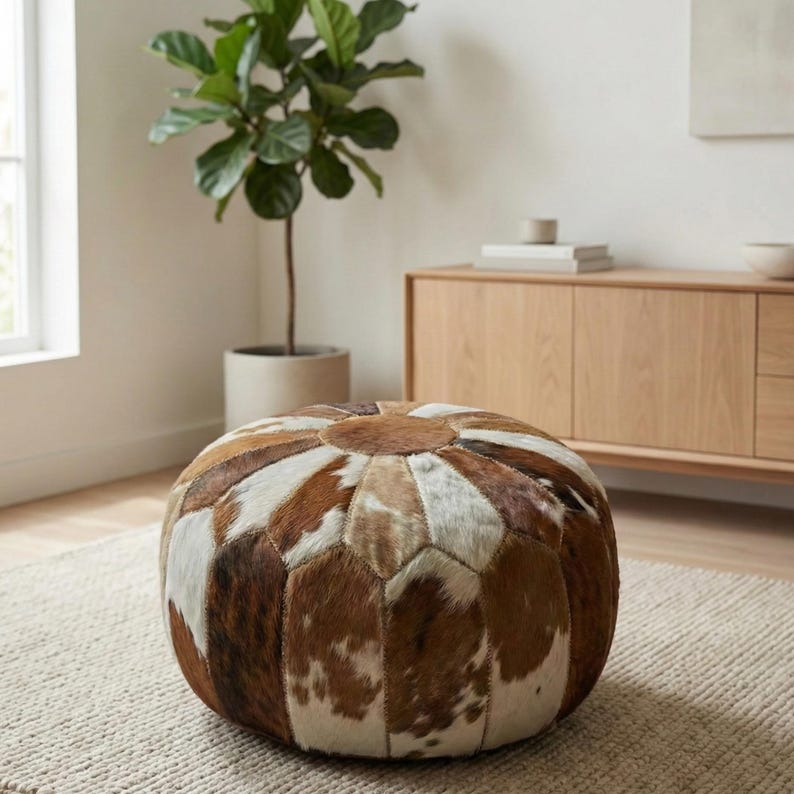 May include: Round ottoman with a patchwork design made from brown, white, and tan cowhide. The ottoman is placed on a neutral-colored rug in a well-lit room. The ottoman is a decorative piece of furniture.