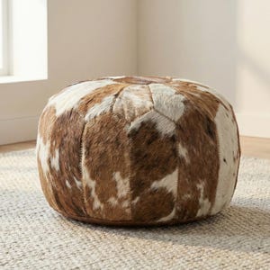 May include: A round, brown and white cowhide ottoman. The ottoman is made of stitched leather patches in shades of brown and white. It sits on a neutral-colored rug and is a decorative piece of furniture.