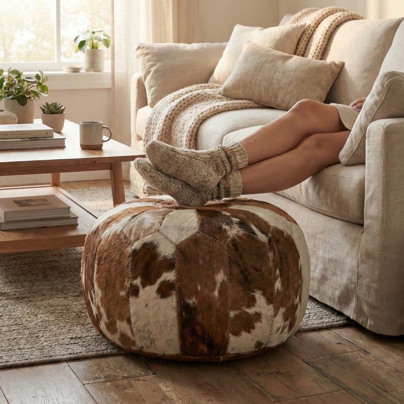 May include: A round, brown and white cowhide ottoman with a patchwork design. The ottoman is in front of a beige sofa with a person relaxing on it. The person is wearing beige socks. A wooden coffee table and plants are in the background.