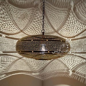 Puede incluir: Una lámpara colgante decorativa con un intrincado diseño perforado. La lámpara está suspendida por una cadena y proyecta una luz estampada sobre la superficie circundante. El color de la lámpara es dorado cálido.