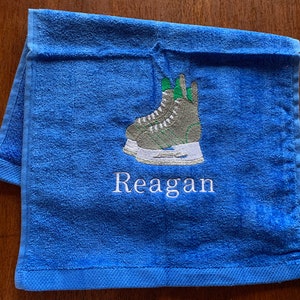 May include: Blue towel with a white embroidered design of a pair of ice skates and the name "Reagan".