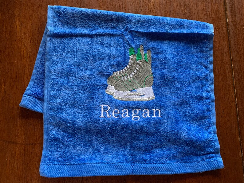May include: Blue towel with a white embroidered design of a pair of ice skates and the name "Reagan".