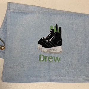 May include: A light blue towel with a green and black embroidered image of ice skates and the name "Drew" in green embroidery.