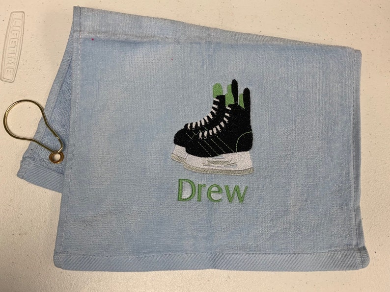 May include: A light blue towel with a green and black embroidered image of ice skates and the name "Drew" in green embroidery.