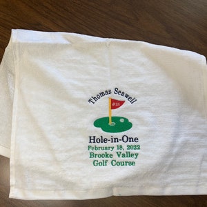 May include: White golf towel with embroidered text and a golf-themed design. The design includes a green golf hole with a flag and a golf ball. Text reads "Thomas Seawell #18 Hole-in-One February 18, 2022 Brooke Valley Golf Course."