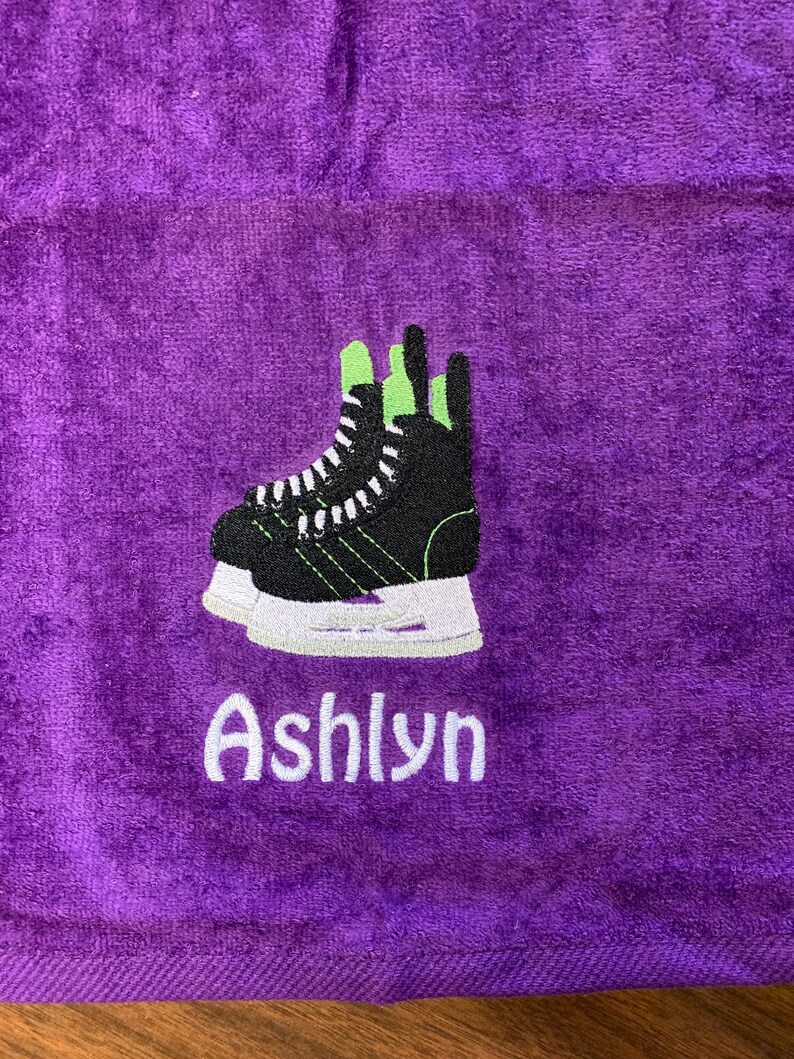May include: Purple towel with a black and white hockey skate embroidered on it. The name "Ashlyn" is embroidered below the skate in white.