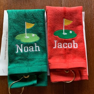 K&ouml;nnte beinhalten: Zwei Golfhandt&uuml;cher, eines gr&uuml;n und eines rot, jeweils mit einem Golfplatzdesign und einem Namen bestickt. Das gr&uuml;ne Handtuch tr&auml;gt den Namen "Noah" und das rote "Jacob". Beide Handt&uuml;cher haben einen goldfarbenen Ring.