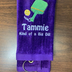May include: Purple towel with a green and yellow pickleball paddle and ball design. The towel has the text "Tammie Kind of a Big Dill" embroidered on it.