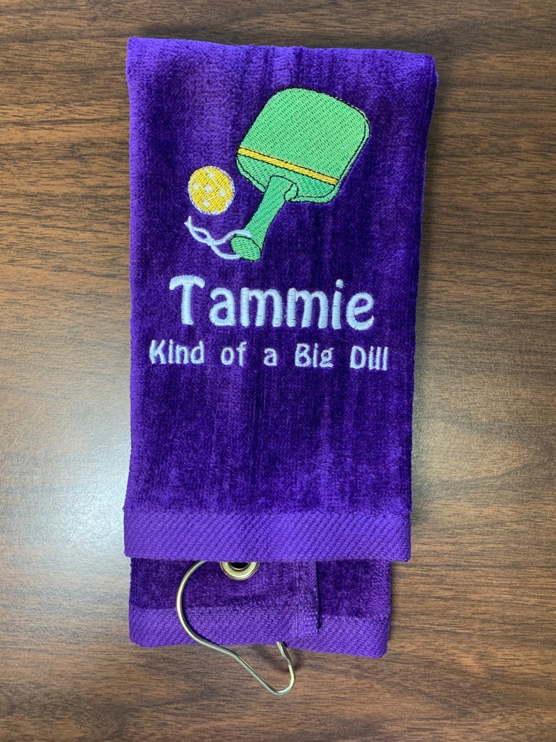 May include: Purple towel with a green and yellow pickleball paddle and ball design. The towel has the text "Tammie Kind of a Big Dill" embroidered on it.