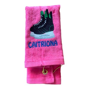 May include: Pink towel with a black and green ice skate embroidered on it. The towel has a loop for hanging and the name "CAITRIONA" is embroidered below the skate.