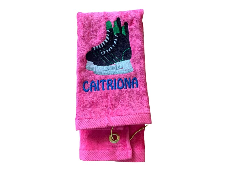 May include: Pink towel with a black and green ice skate embroidered on it. The towel has a loop for hanging and the name "CAITRIONA" is embroidered below the skate.