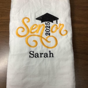 May include: White towel with gold embroidery that says "Senior 2025" with a black graduation cap above the text. The name "Sarah" is embroidered below the text.