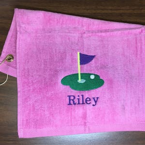 K&ouml;nnte beinhalten: Rosa Golfhandtuch mit einem goldfarbenen Metallring. Das Handtuch zeigt ein gesticktes Golfgr&uuml;n mit einer lila Fahne, einem wei&szlig;en Golfball und dem Namen "Riley" in Blau.