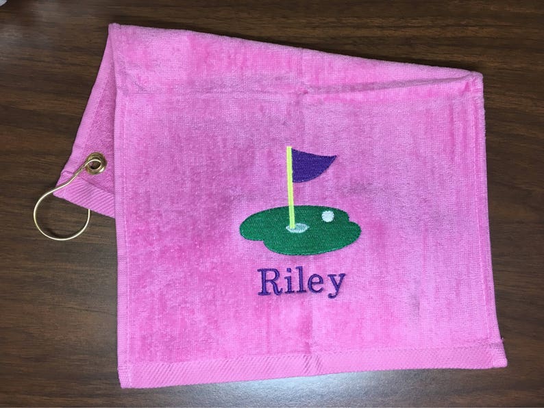 K&ouml;nnte beinhalten: Rosa Golfhandtuch mit einem goldfarbenen Metallring. Das Handtuch zeigt ein gesticktes Golfgr&uuml;n mit einer lila Fahne, einem wei&szlig;en Golfball und dem Namen "Riley" in Blau.
