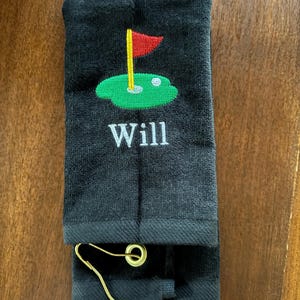 K&ouml;nnte beinhalten: Schwarzes Golfhandtuch mit gestickter gr&uuml;ner Golfplatzszene, roter Fahne und dem Namen "Will" in Wei&szlig;. Enth&auml;lt einen goldfarbenen Metallclip zur Befestigung.