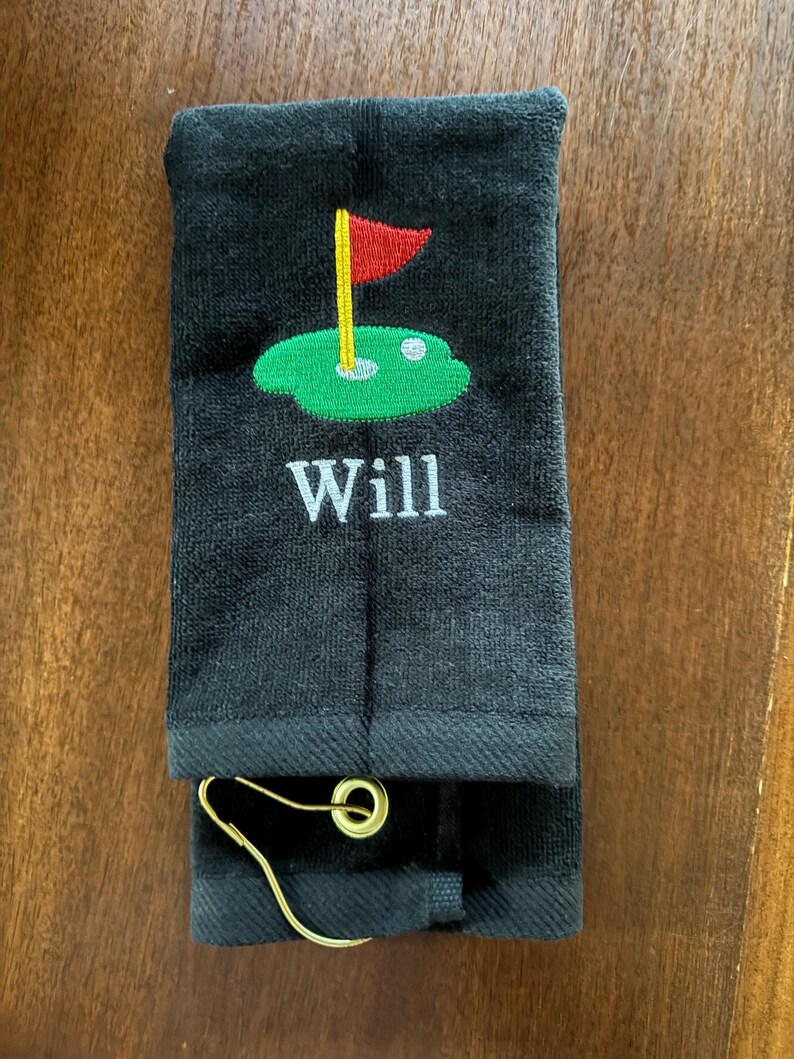 K&ouml;nnte beinhalten: Schwarzes Golfhandtuch mit gestickter gr&uuml;ner Golfplatzszene, roter Fahne und dem Namen "Will" in Wei&szlig;. Enth&auml;lt einen goldfarbenen Metallclip zur Befestigung.