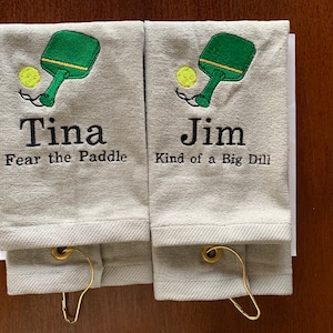 May include: Two white towels with embroidered images of a green paddleball paddle and yellow ball. The towels have personalized names "Tina" and "Jim" with humorous sayings below. "Fear the Paddle" and "Kind of a Big Dill".