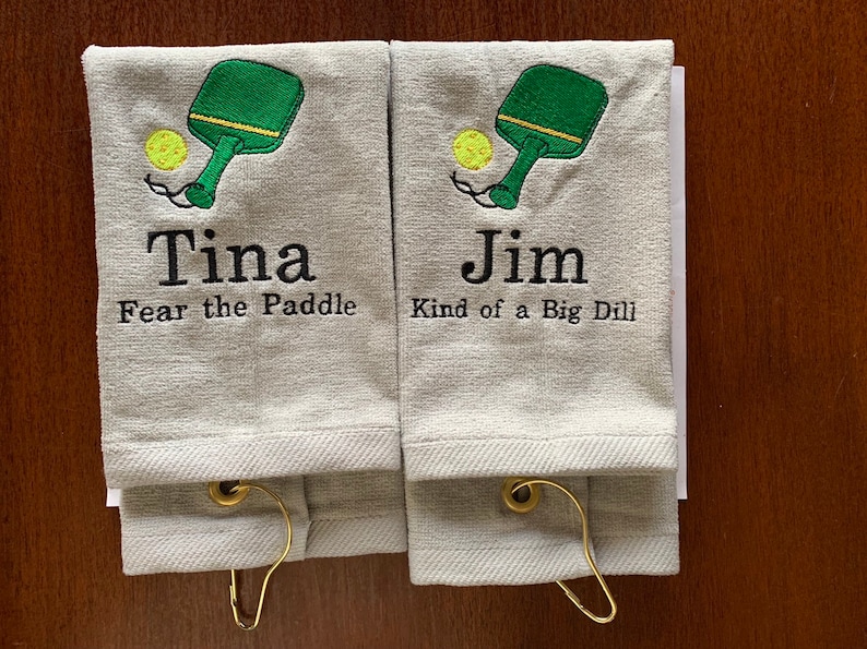 May include: Two white towels with embroidered images of a green paddleball paddle and yellow ball. The towels have personalized names "Tina" and "Jim" with humorous sayings below. "Fear the Paddle" and "Kind of a Big Dill".