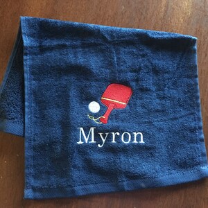 May include: A dark blue towel with a white embroidered name "Myron" and a red and white embroidered ping pong paddle and ball.