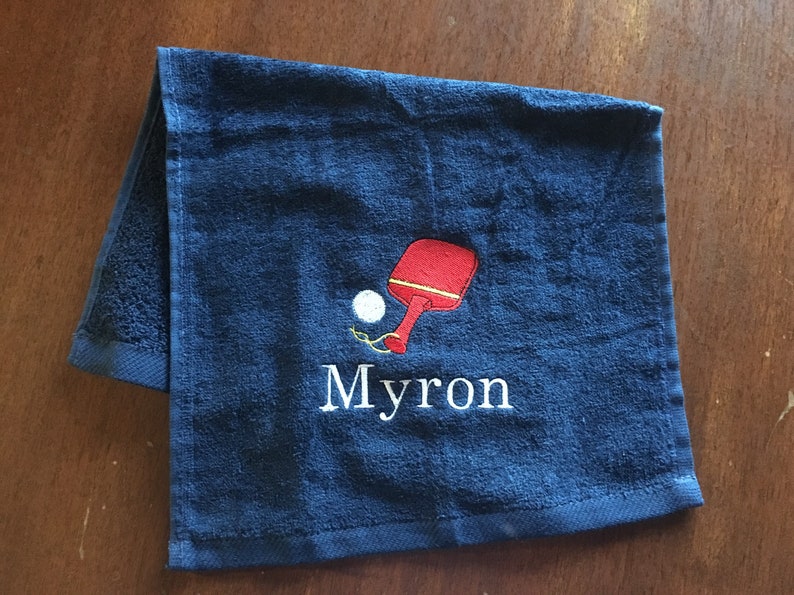 May include: A dark blue towel with a white embroidered name "Myron" and a red and white embroidered ping pong paddle and ball.