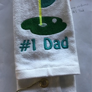 K&ouml;nnte beinhalten: Wei&szlig;es Golfhandtuch mit gesticktem gr&uuml;nem Golfplatzdesign, gelber Fahne und dem Text "#1 Dad" in T&uuml;rkis. Das Handtuch ist gefaltet und hat eine goldfarbene Metallschlaufe.