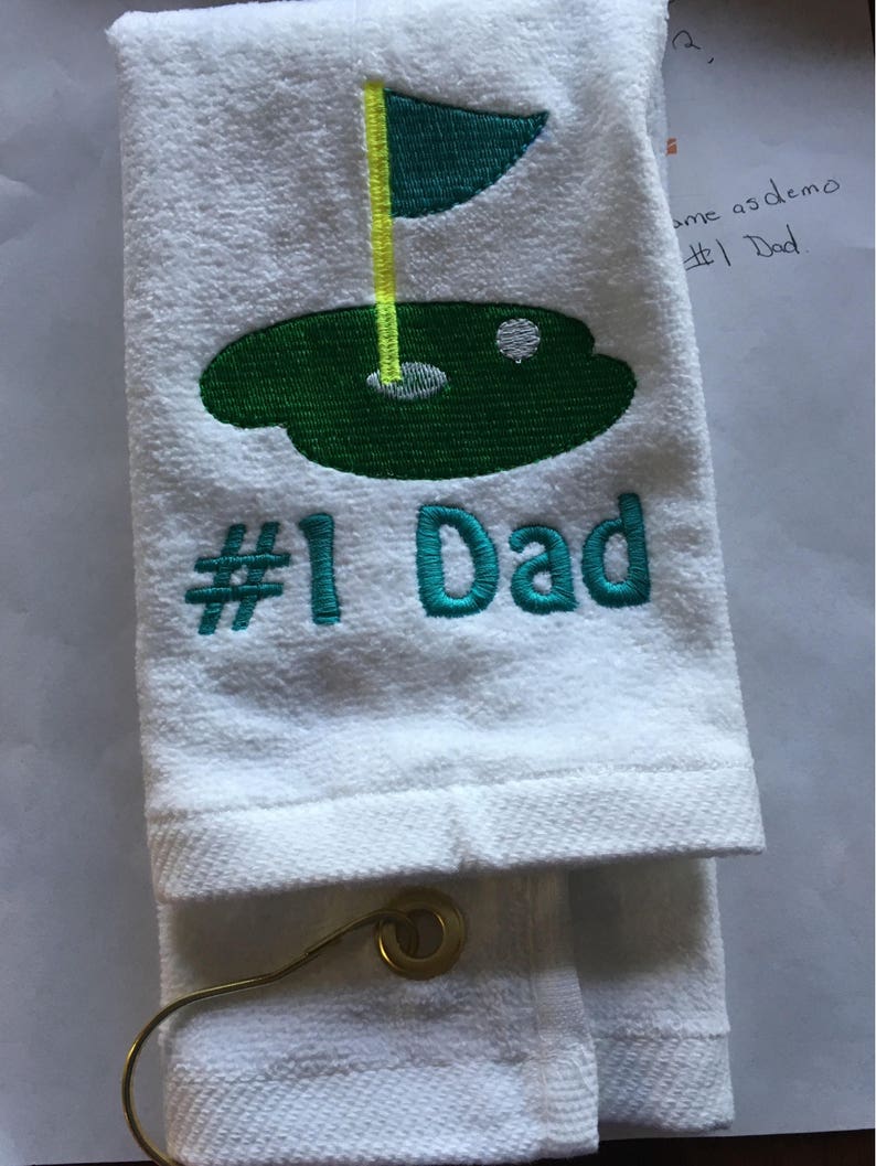 K&ouml;nnte beinhalten: Wei&szlig;es Golfhandtuch mit gesticktem gr&uuml;nem Golfplatzdesign, gelber Fahne und dem Text "#1 Dad" in T&uuml;rkis. Das Handtuch ist gefaltet und hat eine goldfarbene Metallschlaufe.