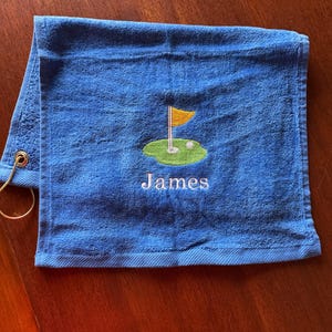 K&ouml;nnte beinhalten: Blaues Golfhandtuch mit goldener Fahne, gr&uuml;nem Putting Green und Golfball-Stickerei. Der Name "James" ist darunter in Wei&szlig; gestickt. Mit einer goldenen &Ouml;se zum Aufh&auml;ngen.