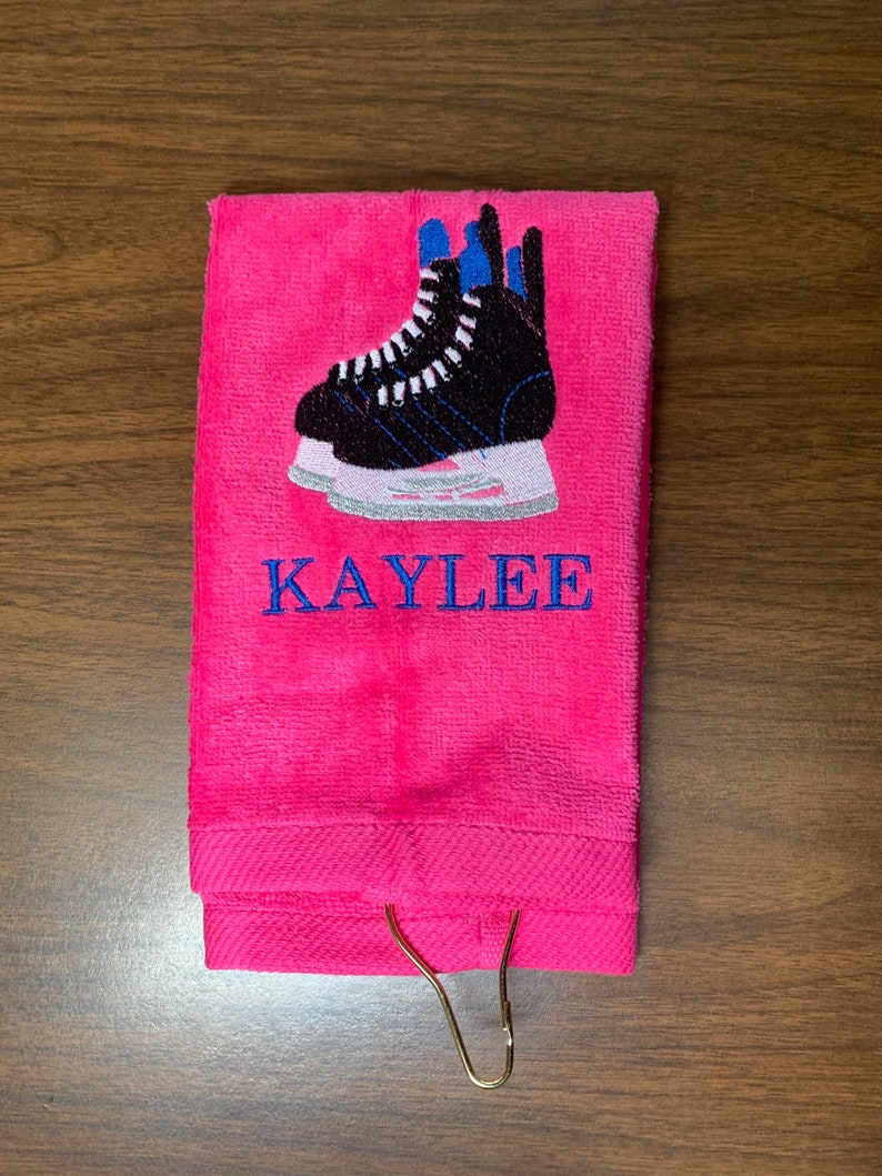 May include: Pink towel with a gold hook, embroidered with a pair of black ice skates and the name "KAYLEE".