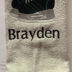 May include: White towel with black embroidery of a green and black ice skate and the name "Brayden".