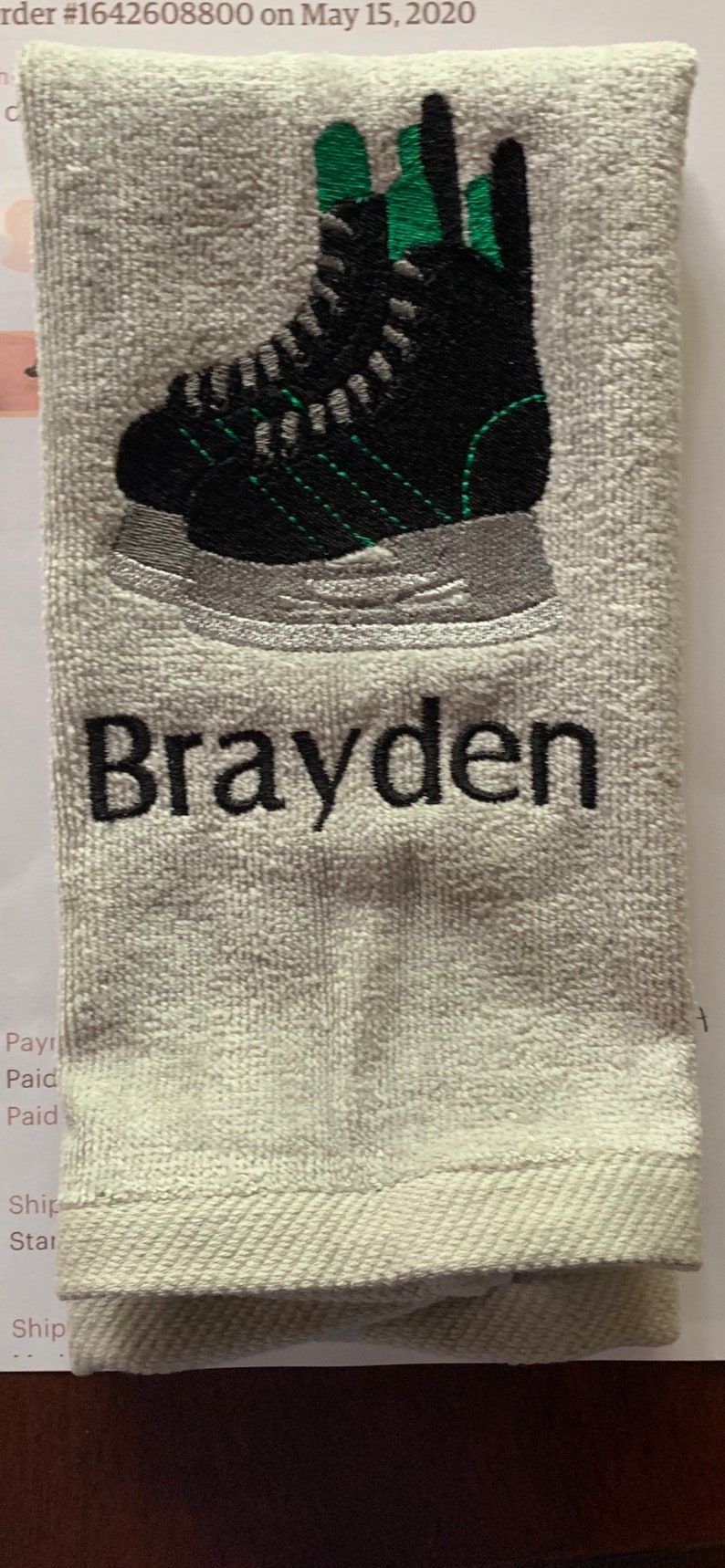 May include: White towel with black embroidery of a green and black ice skate and the name "Brayden".