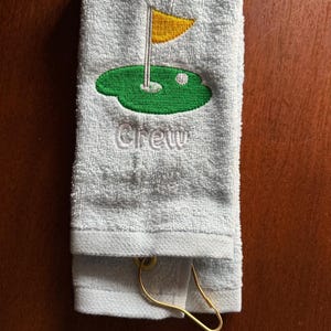 K&ouml;nnte beinhalten: Hellblaues Golfhandtuch mit gesticktem gr&uuml;nem Putting Green, gelber Fahne und wei&szlig;em Golfball. Das Wort "Crew" ist darunter gestickt. Ein goldfarbener Metallclip ist befestigt.