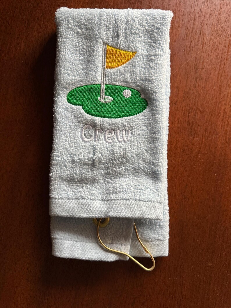 K&ouml;nnte beinhalten: Hellblaues Golfhandtuch mit gesticktem gr&uuml;nem Putting Green, gelber Fahne und wei&szlig;em Golfball. Das Wort "Crew" ist darunter gestickt. Ein goldfarbener Metallclip ist befestigt.